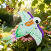 A plush toy resembling a Luna moth with green and purple wings sitting among flowers in natural sunlight.