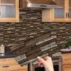 A hand holds a rectangular wooden tile sticker for kitchen backsplash against a textured brown stone wall.