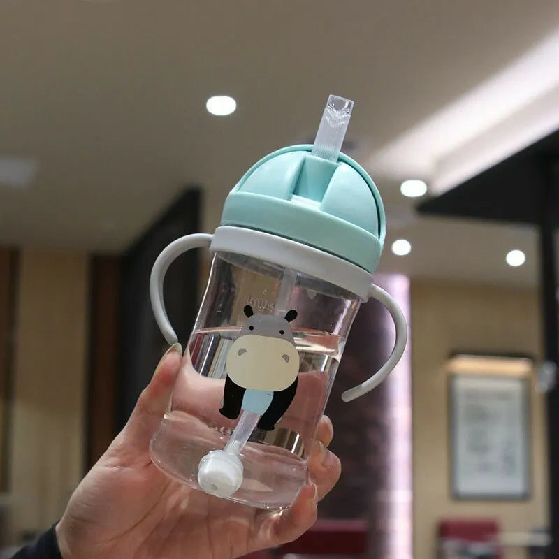 A hand holds a clear sippy cup with a light blue lid and gravity straw, featuring a black bear design.