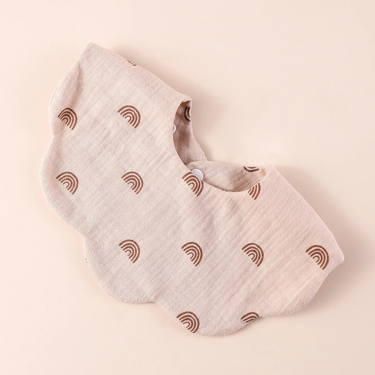 A light beige cotton baby bib with a repeating rainbow pattern shown from above.