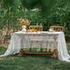 An elegant white lace tablecloth set up outdoors on a wooden table with floral arrangements.