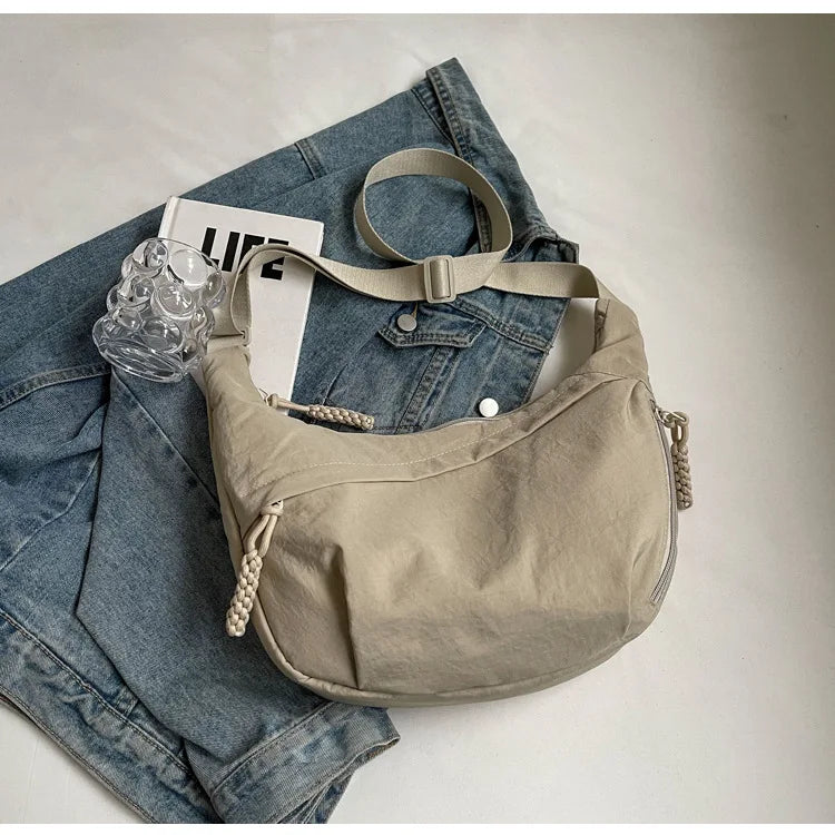 A beige lightweight nylon crossbody bag resting on a pair of blue denim jeans.
