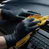 A hand wearing a black nitrile glove is cleaning a black electronic device with a yellow cloth.
