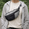 A person wearing a light gray jacket and cream shirt holds an everyday nylon crossbody bag with a black mesh pocket.