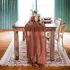 A rustic table set with a terracotta gauze fabric runner on a wooden table and white rug.