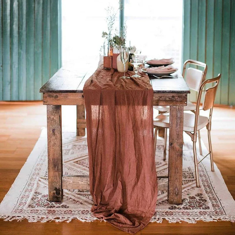 A rustic table set with a terracotta gauze fabric runner on a wooden table and white rug.