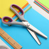 A pair of silver professional pinking shears lying on a blue surface next to colored paper and a wooden ruler.