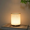 A white fabric desk lamp with a warm glow illuminates a surface next to a green leaf.
