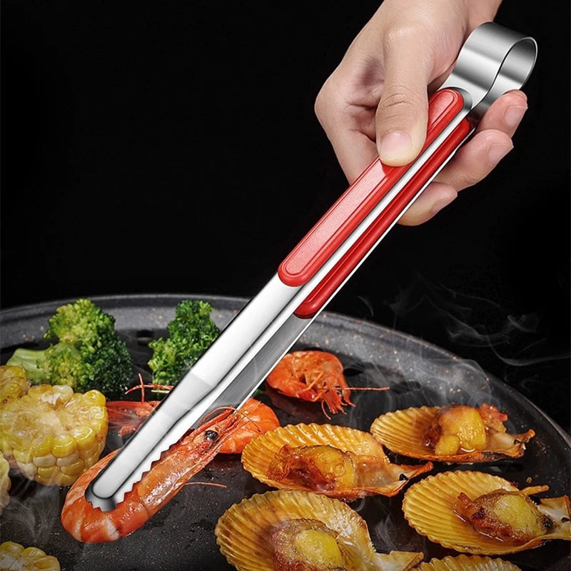 A hand holds a pair of stainless steel BBQ tongs used for grilling and serving seafood on a dark pan.