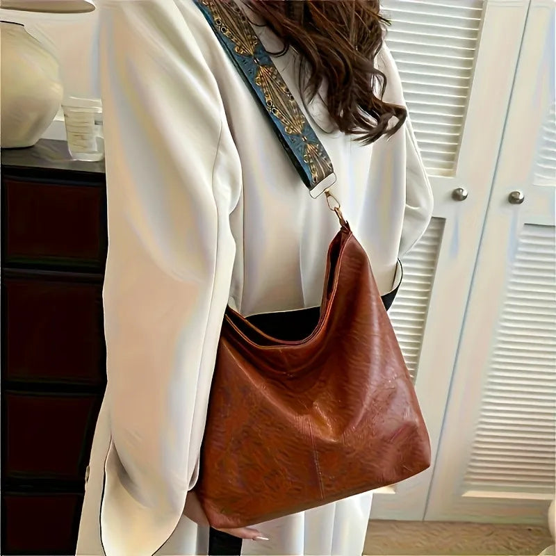 A woman holds a brown leather shoulder bag with golden embroidery against a white blazer and light wooden blinds.