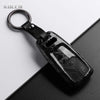 A black carbon fiber style key fob cover with a metal ring attached, shown on a dark surface.