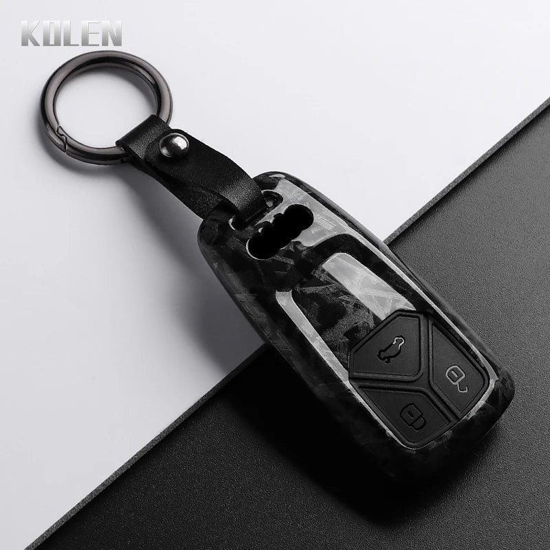 A black carbon fiber style key fob cover with a metal ring attached, shown on a dark surface.