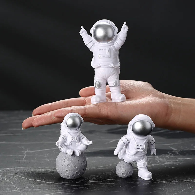 A pair of handcrafted astronaut figurines, one standing on a hand and another resting on a sphere.