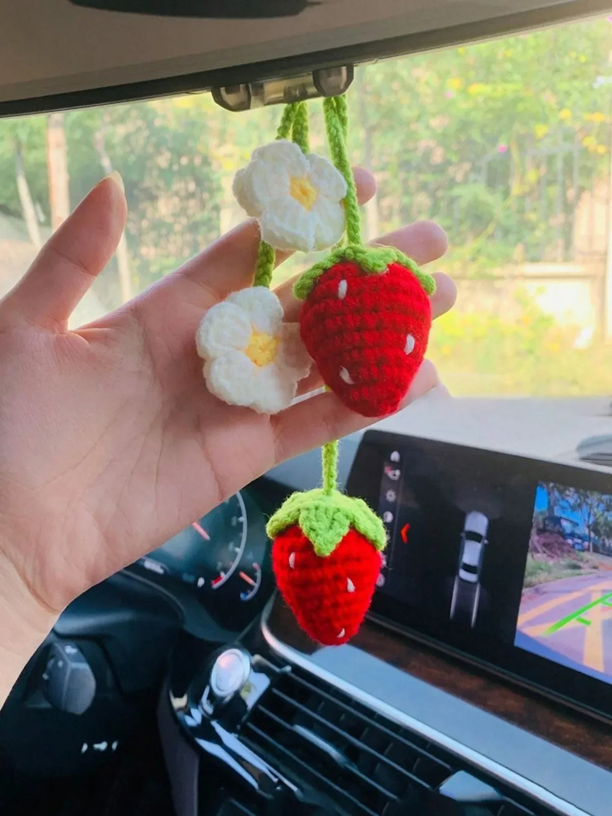 Hand holding a handmade crochet strawberry and flower pendant against a car mirror background