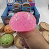 Eine Hand hält ein großes, pinkes Knet-Stress-Spielzeug neben ähnliche Spielzeuge in ihrer Verpackung.
