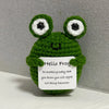 A handmade crochet frog doll with large eyes and a green body sits on a white surface.