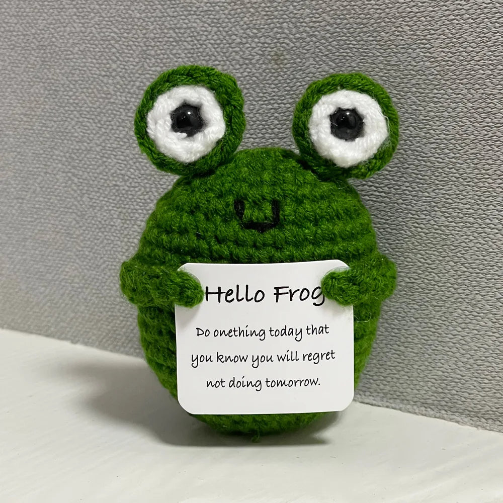 A handmade crochet frog doll with large eyes and a green body sits on a white surface.