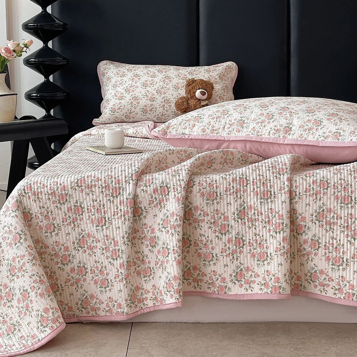 A floral quilted bedspread in pink and cream patterns draped over a dark headboard.