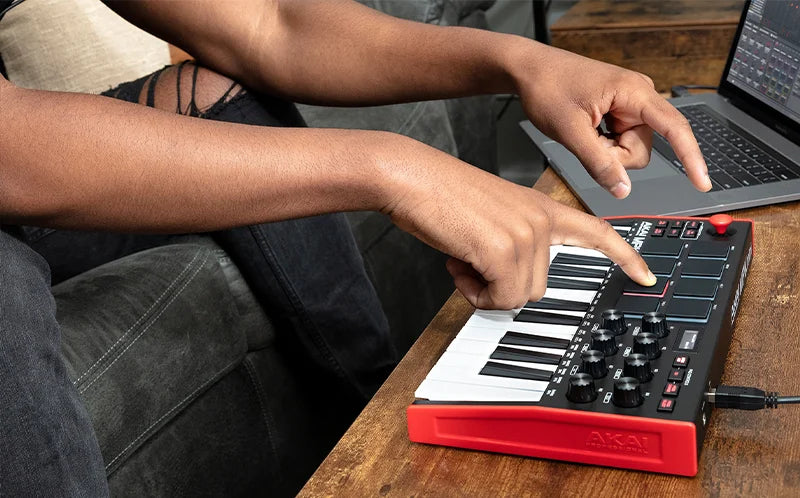 A person's hand is playing an AKAI Professional MPK Mini MK3 MIDI keyboard controller with drum pads and knobs on a wooden ta