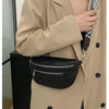 A close-up of a woman wearing a beige trench coat, holding a black multi-pocket PU leather waist bag with an adjustable strap