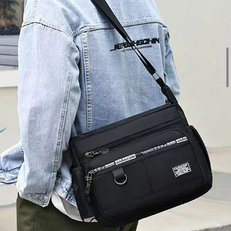 A black men's business crossbody bag with multiple zippered pockets worn over a light blue denim shirt.