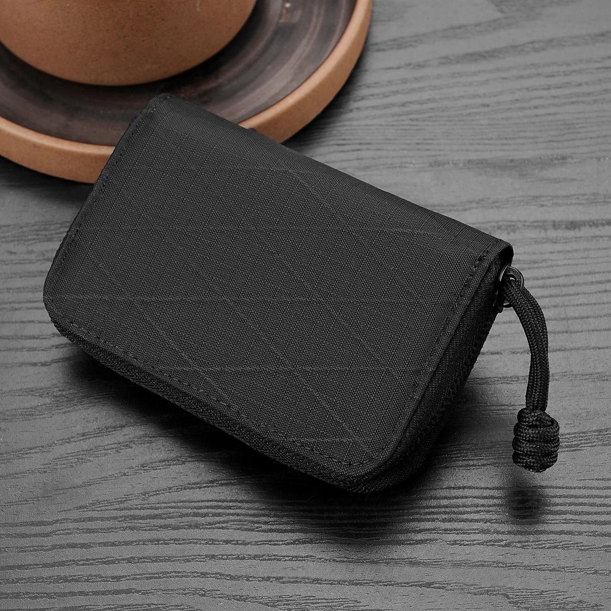 A black compact RFID wallet with a zipper pull is shown resting on a gray wooden surface next to a ceramic plate.
