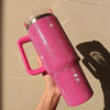 A hand holds a bright pink rhinestone tumbler with a straw and handle against a light background.