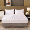 A white fitted mattress protector with an elastic edge resting on a gray upholstered bed base.