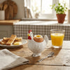 A white ceramic chicken egg cup with a yellow yolk sits on a wooden table next to a glass of orange juice.