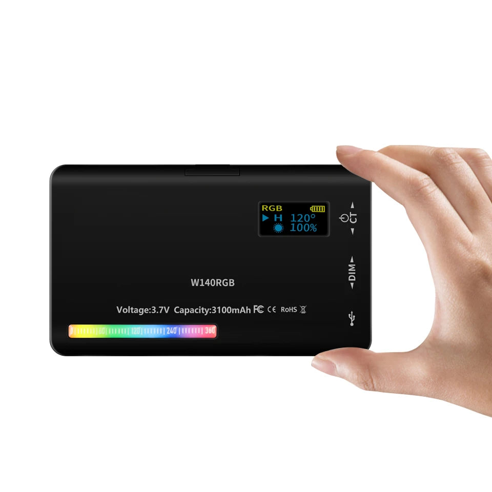 A hand holds a black portable RGB LED video light panel displaying battery and power information.