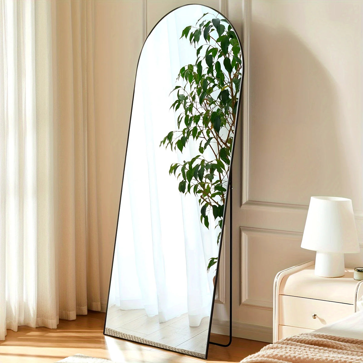 An arched full-length floor mirror with a black aluminum frame reflecting greenery in a bright room.