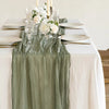 A rustic country boho table setting featuring a sage green cotton gauze table runner with white floral arrangements.