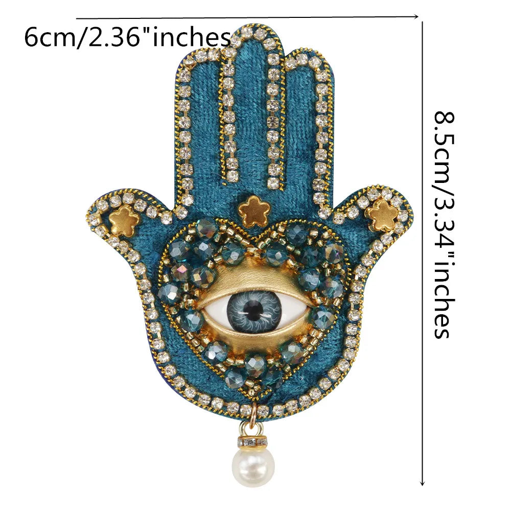 A handprint design featuring blue fabric, gold accents, and a central eye made of sparkling beads and rhinestones.