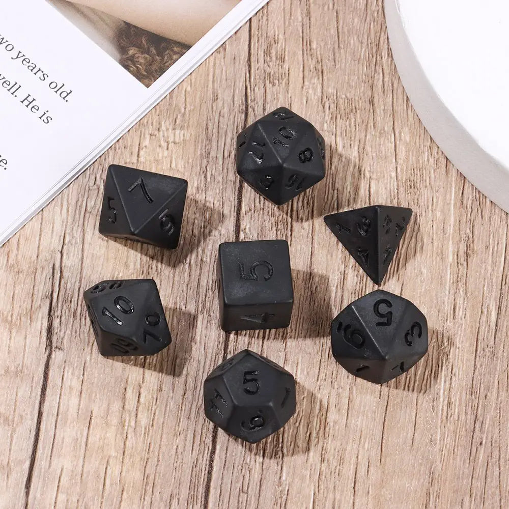 A set of matte black polyhedral dice, including a d7, d10, d12, d20, and d100, scattered on a wooden surface.