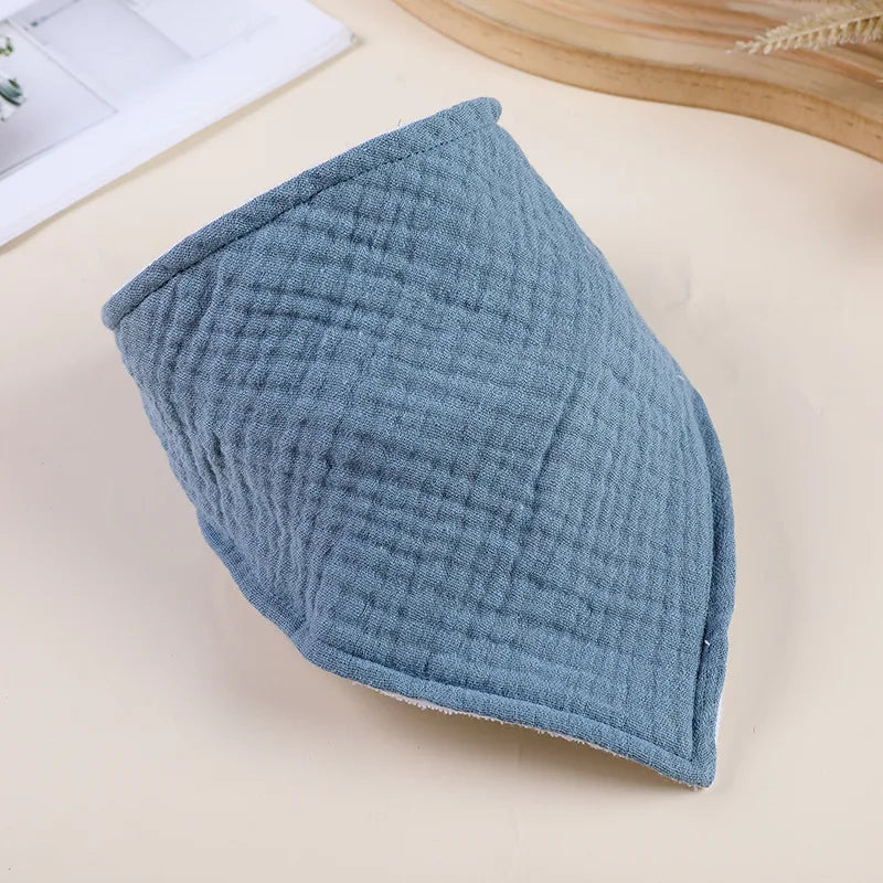 A light blue, textured cotton baby bib is shown angled on a beige surface.