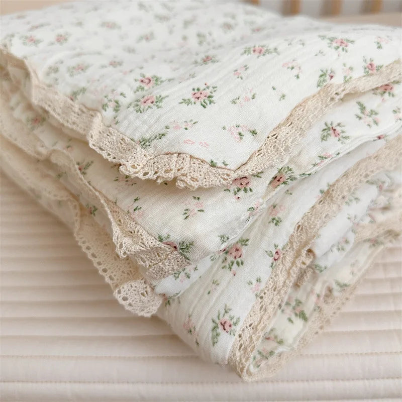 A stack of white floral muslin baby blankets with delicate lace trim, folded on a light pink surface.