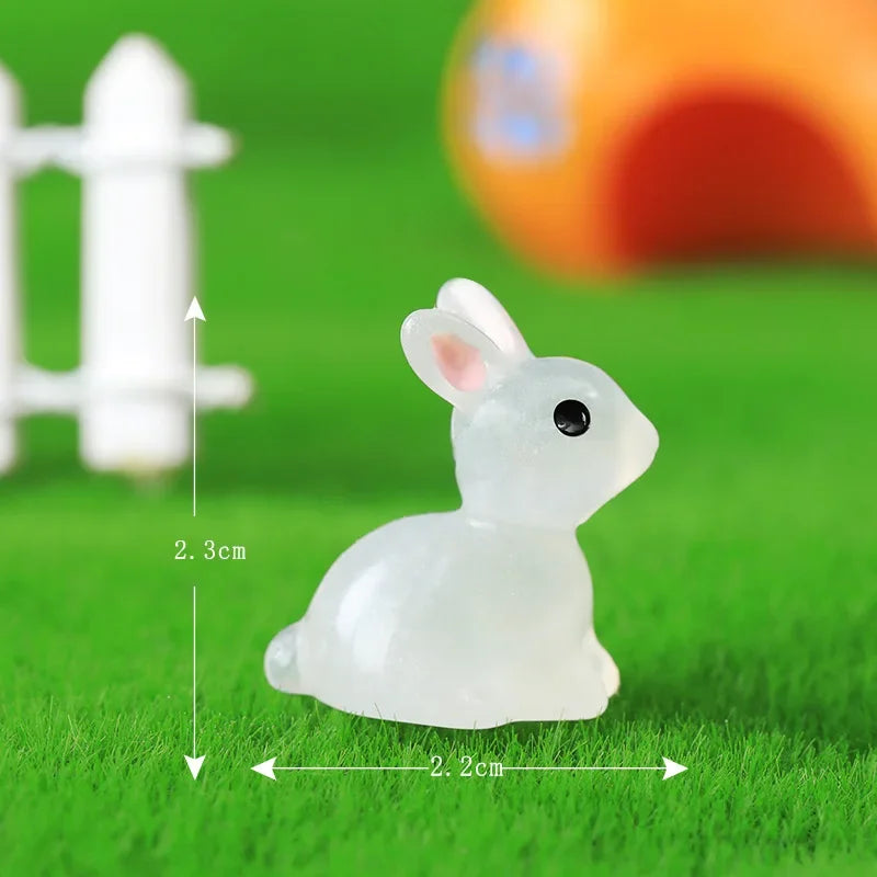 A white resin miniature bunny figurine sitting on green grass with a white fence in the background.