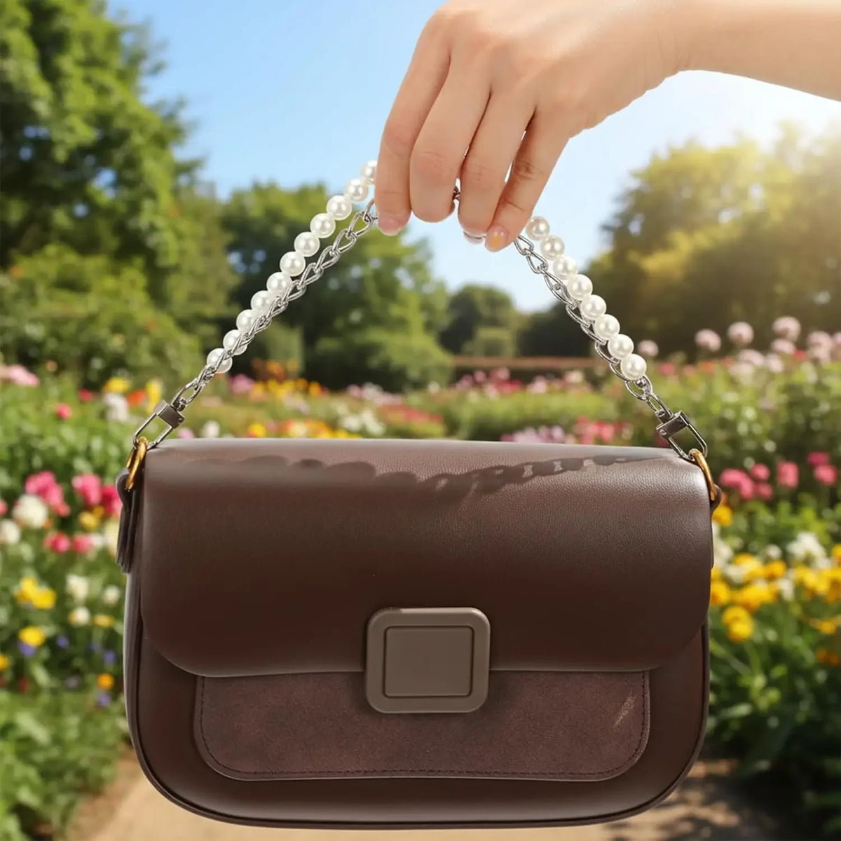 A hand holds an elegant brown purse with a pearl and gold chain in a garden setting.