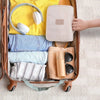 A beige travel makeup bag with a hanging hook is shown inside a brown suitcase, surrounded by folded clothing and cosmetics.