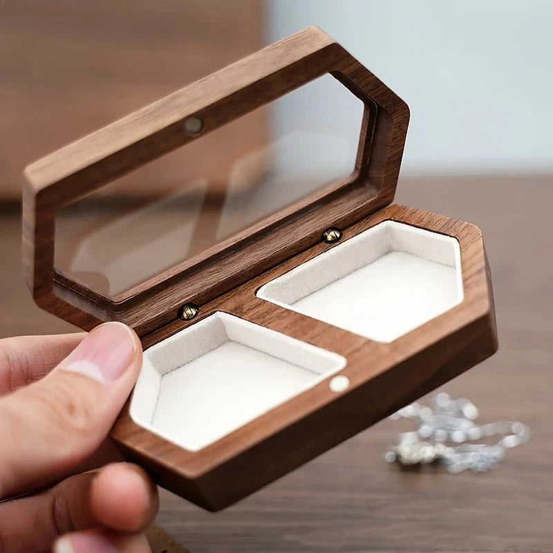 A hand holds an open walnut wood ring box with a clear lid and two compartments, showing the interior velvet lining.