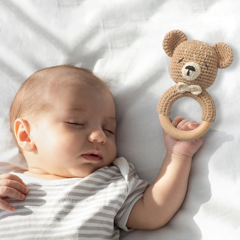 A baby is holding a handmade crochet teddy bear rattle with a natural wood teether ring.