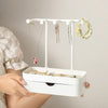 A white elegant jewelry display rack with a drawer and wooden base holding various necklaces and earrings.
