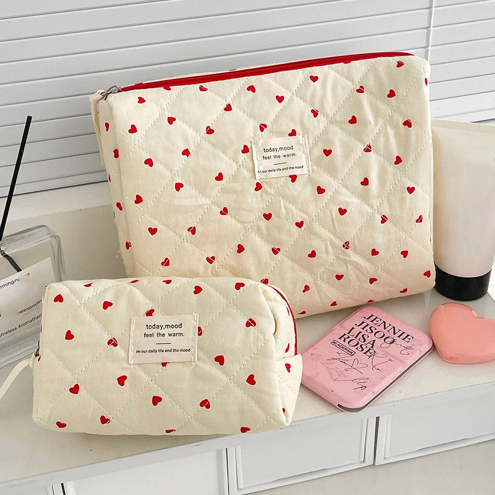 A cream quilted makeup bag with a red piping edge and a repeating pattern of small red hearts, shown next to a pink compact.