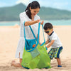 A woman is holding a large mesh green beach tote bag while interacting with a young boy on a sandy beach.