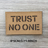 A rectangular tan fabric patch with the text TRUST NO ONE printed in black block letters on a weathered wooden surface.