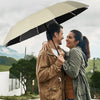 A couple walks outdoors under a large beige umbrella protecting them from the rain.
