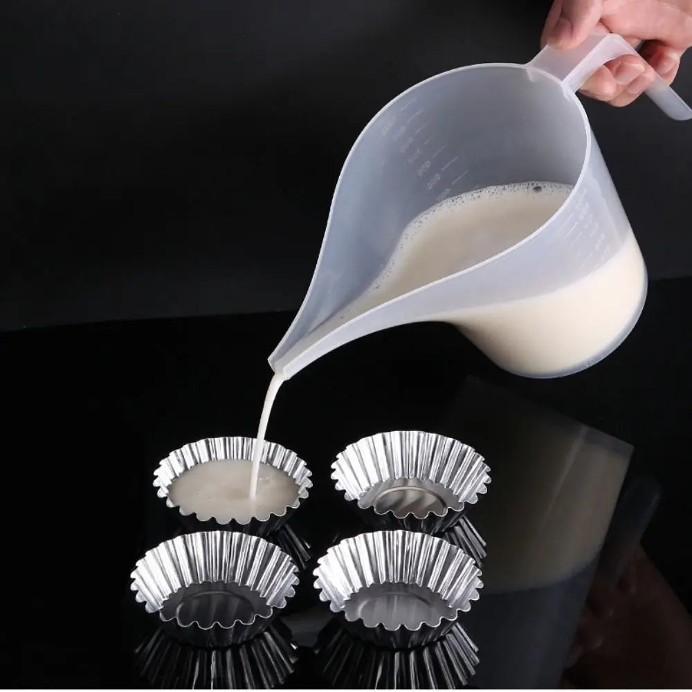 A hand pours white liquid from a plastic measuring cup with a long spout into several small metal measuring cups.