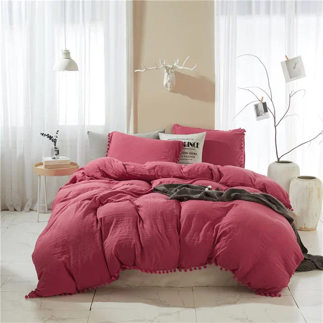 A pink duvet cover set with pom tassels is displayed on a bed in a bright room.