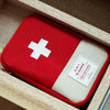 A red and white compact travel medicine organizer bag is placed inside a light wood box.