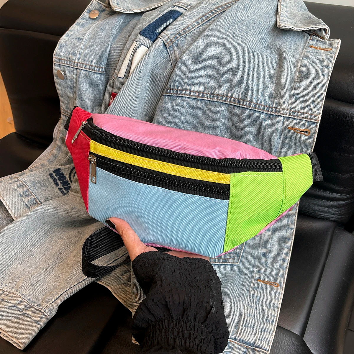 A hand holds a colorful nylon crossbody waist pack with pink, yellow, and blue sections against a denim jacket.